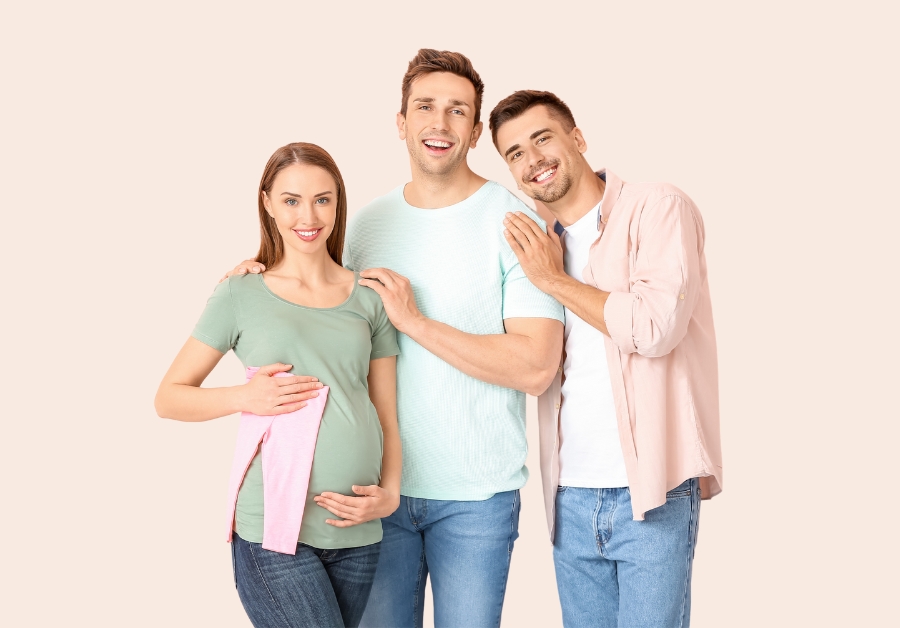 Three people, including a pregnant surrogate, standing together, smiling and looking happy.