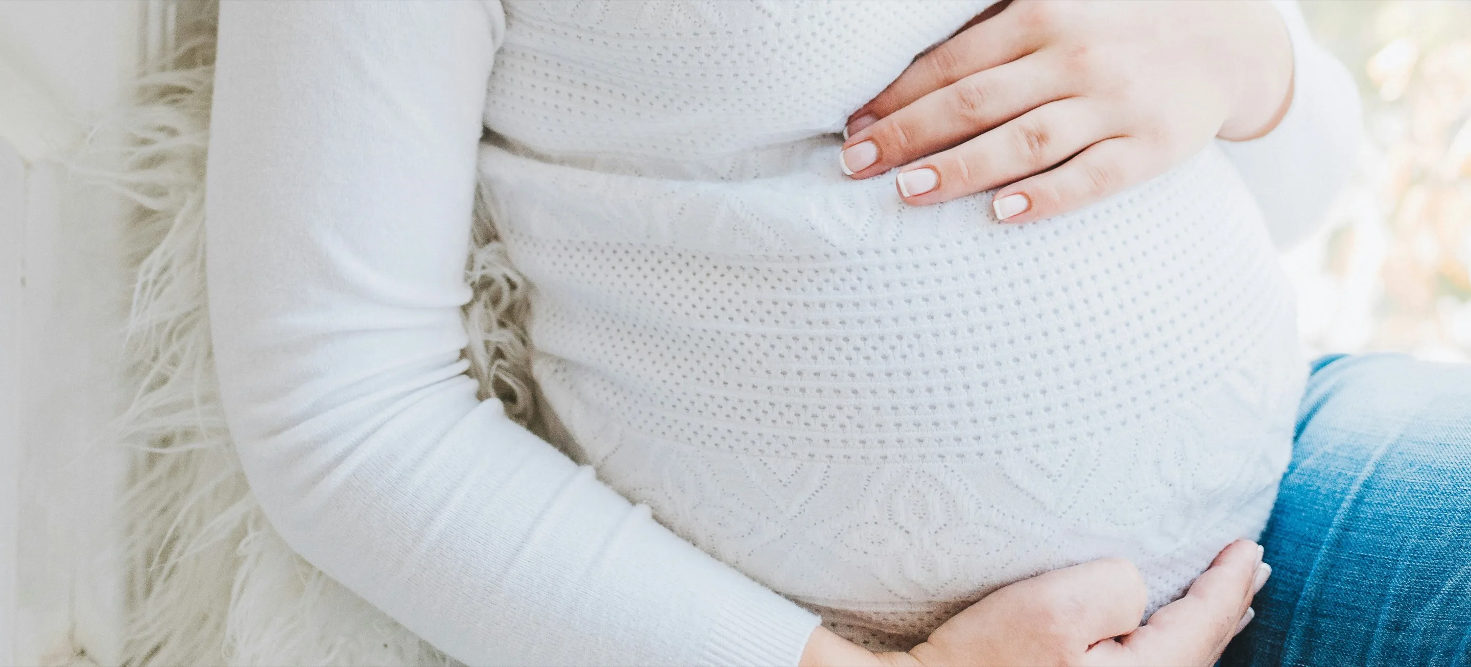 Pregnant woman in a white sweater resting both hands on her belly