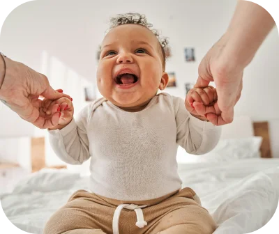 A smiling baby sitting up and reaching for an adult's hand