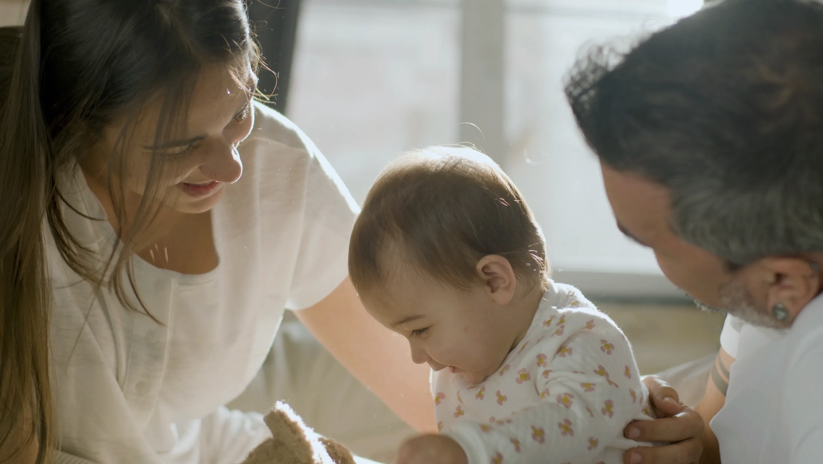 Mother and father leaning in close to their smiling baby on a sunlit bed